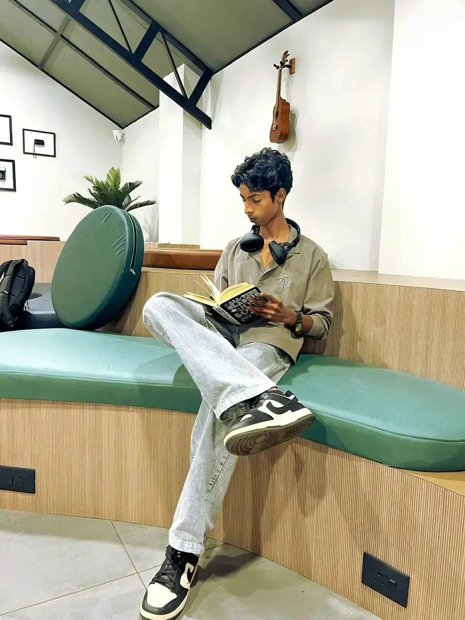 Photo of a young male sitting on a light blue bench, reading a book with headphones around his neck, in a modern, casual setting.