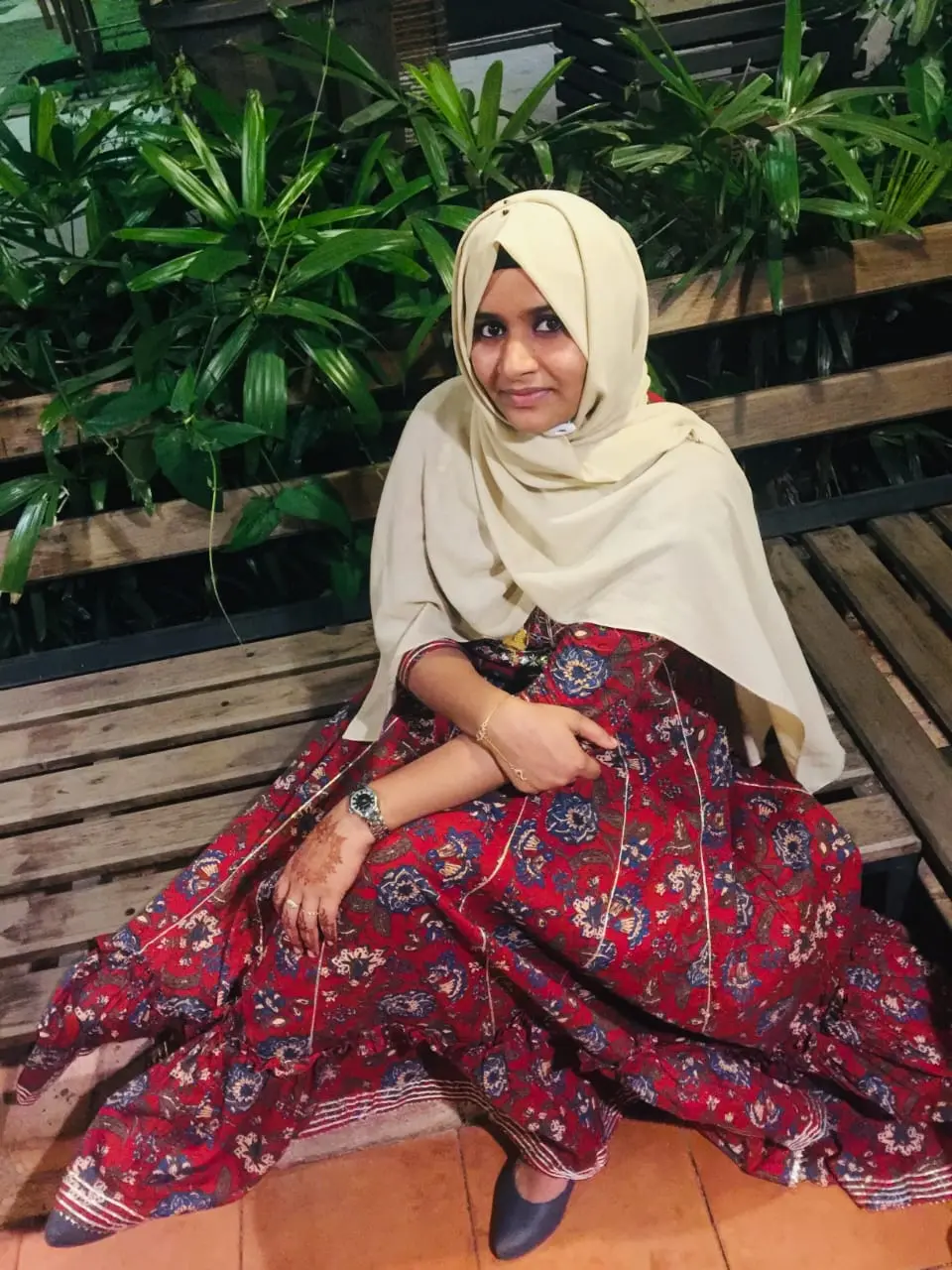 Photograph of a client wearing a red floral dress and light-colored hijab, sitting on a wooden bench outdoors at night.